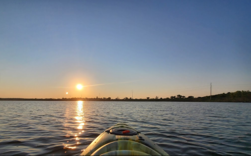 A kayak on calm water at sunset, with the sun reflecting on the surface and a clear sky in the background.