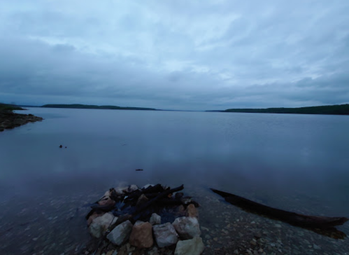 A calm lake at dusk, with a rocky shore and a few scattered logs under a cloudy sky.