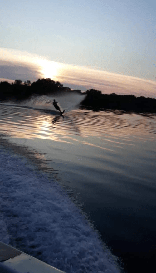 A person water skiing on a calm lake at sunset, with reflections on the water and a gentle spray behind them.