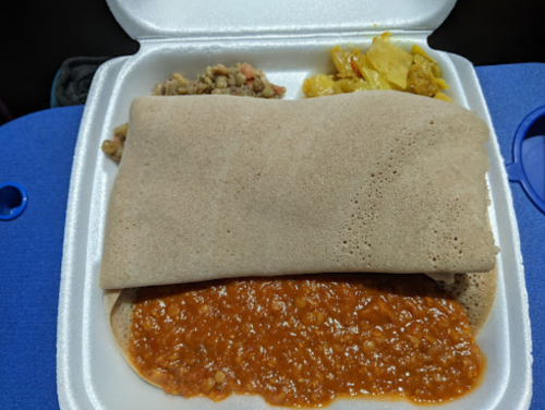 A takeout container with injera topped with lentil stew, accompanied by a side of vegetables and lentils.