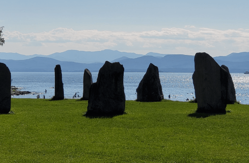 A row of standing stones on a grassy shore, overlooking a calm lake and distant mountains under a clear blue sky.