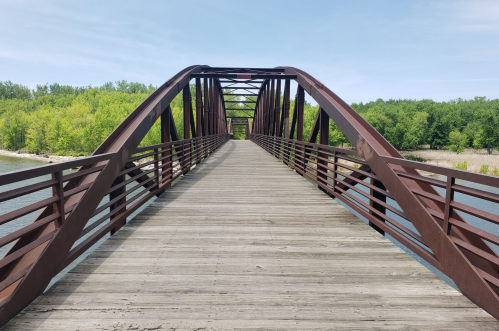 A wooden bridge with metal arches spans a calm river, surrounded by lush green trees under a clear blue sky.