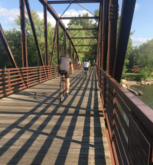 Two cyclists ride on a wooden bridge with metal beams, surrounded by trees and a river in the background.