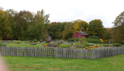 A red house surrounded by colorful trees and a garden enclosed by a wooden fence on a cloudy day.