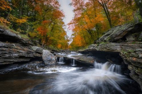 A serene river flows through a rocky landscape, surrounded by vibrant autumn foliage in warm hues.