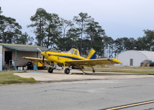 A yellow crop-dusting airplane parked on a tarmac near a hangar, surrounded by trees and a cloudy sky.