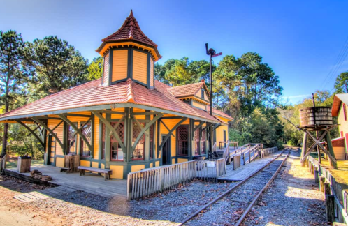 Historic train station with a distinctive tower, surrounded by trees and a railway track under a clear blue sky.