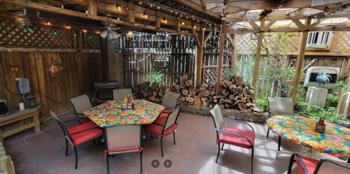 A cozy outdoor patio with colorful tablecloths, wooden walls, and stacked firewood in the background.