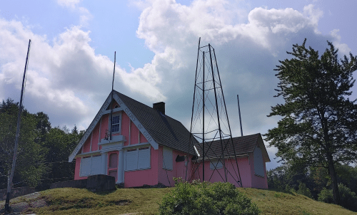 A pink house with boarded windows stands on a hill, next to a tall metal tower under a cloudy sky.