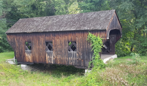 A rustic wooden covered bridge with diamond-shaped windows, surrounded by greenery and grass.
