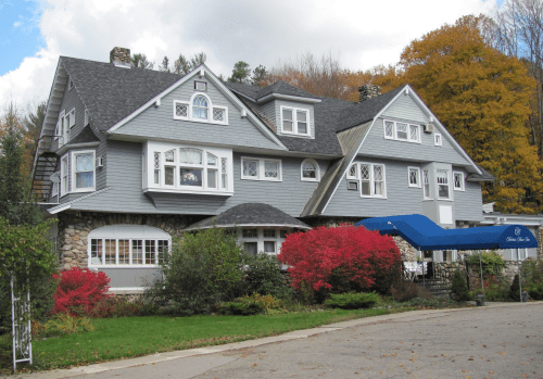 A large gray house with multiple gables, surrounded by colorful shrubs and trees, featuring a blue awning at the entrance.
