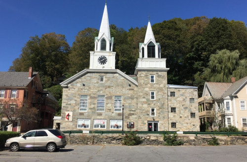 Historic stone church with twin spires, surrounded by trees and nearby buildings under a clear blue sky.