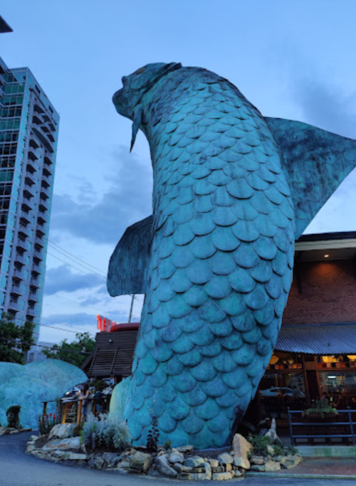 A large, turquoise fish sculpture stands outside a building against a twilight sky.