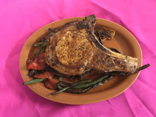 A grilled pork chop served on a plate with roasted green beans and tomatoes, set against a pink background.