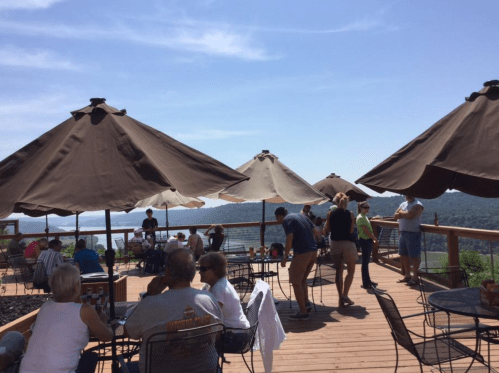 A lively outdoor deck with umbrellas, people dining, and scenic views of hills and water under a clear blue sky.