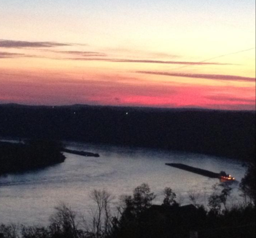 A serene river at sunset, with vibrant pink and purple skies reflecting on the water and a barge navigating the river.