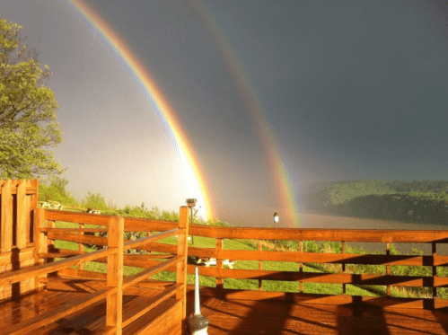 A vibrant double rainbow arcs over a lush landscape, seen from a wooden deck under a stormy sky.