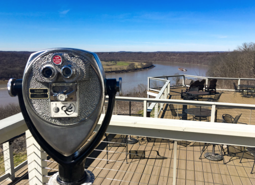 A coin-operated telescope overlooks a river and scenic landscape from a deck with tables and chairs.