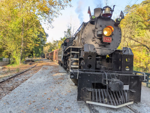 A vintage steam locomotive on a gravel track surrounded by trees in autumn colors.