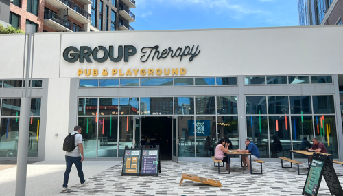Exterior of "Group Therapy Pub & Playground" with people seated outside and a passerby walking by.