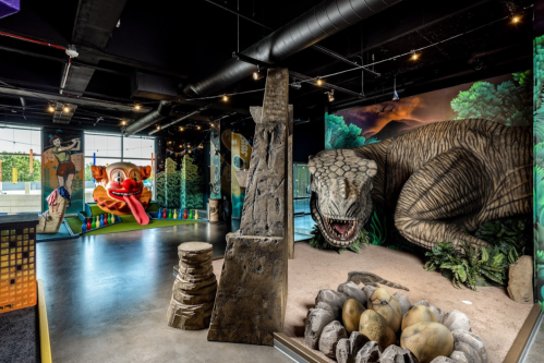 A vibrant indoor play area featuring a dinosaur mural, playful clown art, and rock formations with dinosaur eggs.
