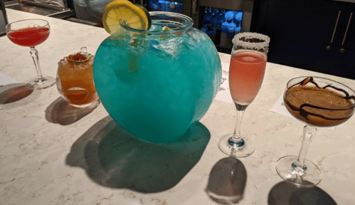 A colorful assortment of cocktails, including a large blue drink in a fishbowl, on a marble bar counter.