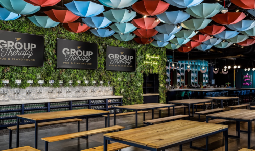 A vibrant pub with colorful umbrella decor, greenery, and wooden tables, featuring a bar and multiple drink taps.