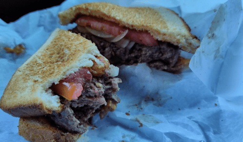 A close-up of a half-eaten sandwich with beef, tomatoes, and onions, served on toasted bread and wrapped in paper.