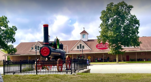 A vintage steam locomotive stands in front of an antique marketplace building under a cloudy sky.
