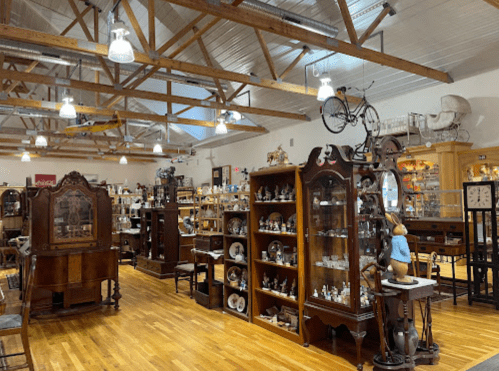 A spacious antique shop featuring wooden furniture, display cases, and various collectibles under a wooden-beamed ceiling.