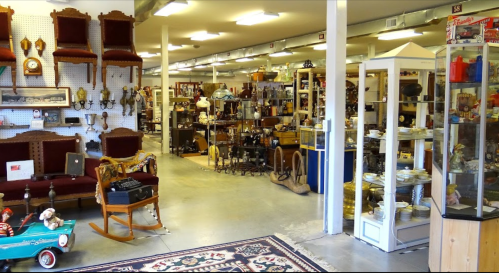 A spacious antique shop filled with vintage furniture, collectibles, and decorative items, with a colorful rug in the foreground.