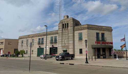 Historic fire museum building with a tall tower, surrounded by flags and parked cars under a cloudy sky.