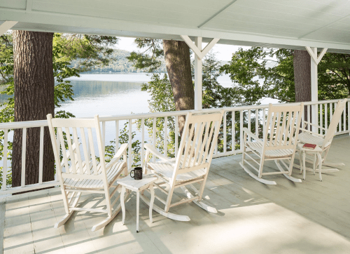 A serene porch with white rocking chairs overlooking a calm lake surrounded by trees.