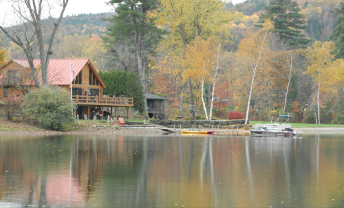 A serene lakeside scene featuring a wooden cabin, autumn trees, and boats on calm water.