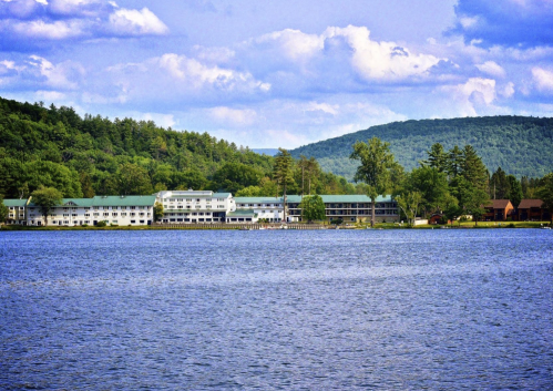 A serene lakeside view featuring a hotel surrounded by lush green hills and a clear blue sky.