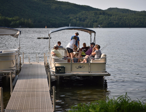 A group of people fishing from a boat on a lake, with a dock and lush greenery in the background.