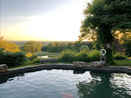 A serene view of a pool at sunset, surrounded by lush greenery and rolling hills in the background.