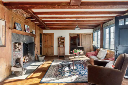 Cozy living room with wooden beams, a fireplace, a brown sofa, and a colorful rug, featuring natural light from windows.