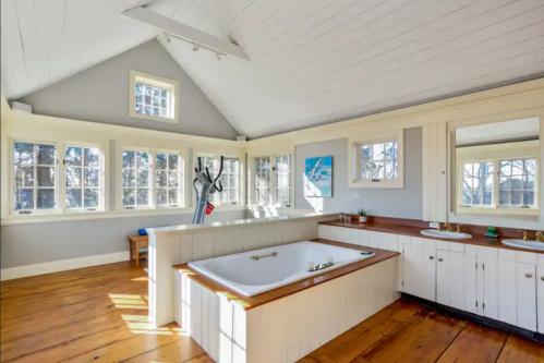Bright, spacious bathroom with a freestanding tub, wooden floors, large windows, and a mirror.