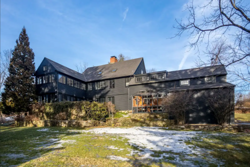 A large, dark-colored house with multiple windows, surrounded by greenery and a clear blue sky.