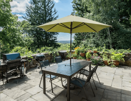 A patio with a table, chairs, and an umbrella, surrounded by greenery and potted plants under a blue sky.