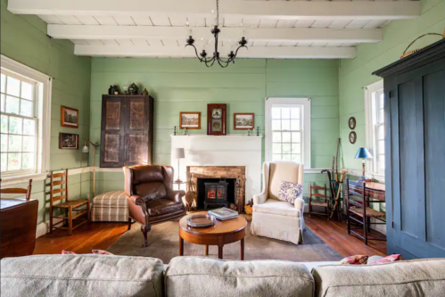 Cozy living room with green walls, wooden beams, a fireplace, and comfortable seating. Warm and inviting atmosphere.