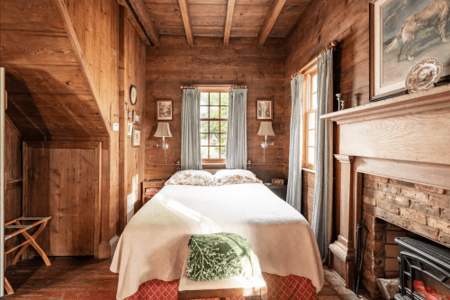 Cozy wooden bedroom with a double bed, fireplace, and natural light from a window, featuring rustic decor.