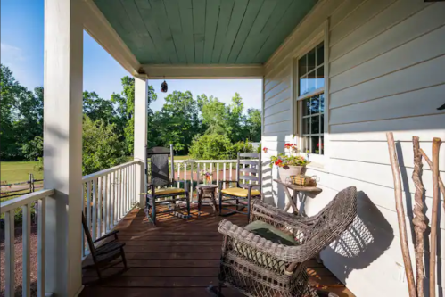 A cozy porch with rocking chairs, a small table, and potted plants, surrounded by greenery and sunlight.
