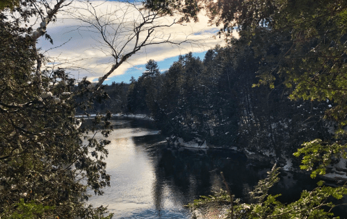 A serene river flows through a snowy landscape, surrounded by trees under a partly cloudy sky.