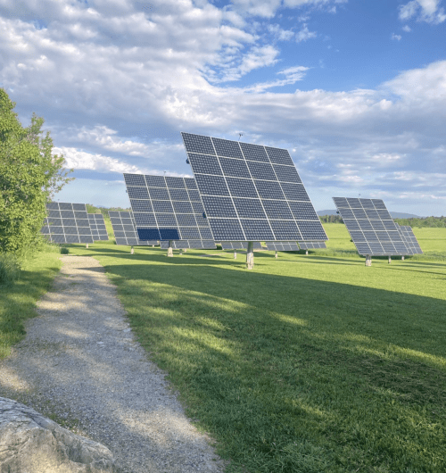 A row of solar panels on a grassy field under a partly cloudy sky, with a gravel path in the foreground.