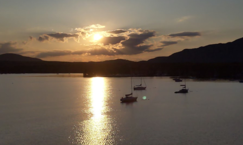 Sunset over a calm lake with silhouettes of sailboats and mountains in the background, reflecting golden light on the water.