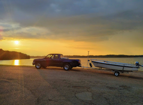 A pickup truck towing a boat at sunset by a calm lake, with a colorful sky and trees in the background.