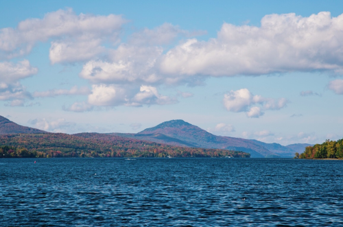 A serene lake surrounded by mountains and colorful autumn foliage under a blue sky with fluffy clouds.