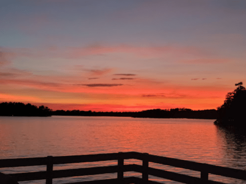 A serene lake at sunset, with vibrant orange and pink hues reflecting on the water and silhouettes of trees in the background.